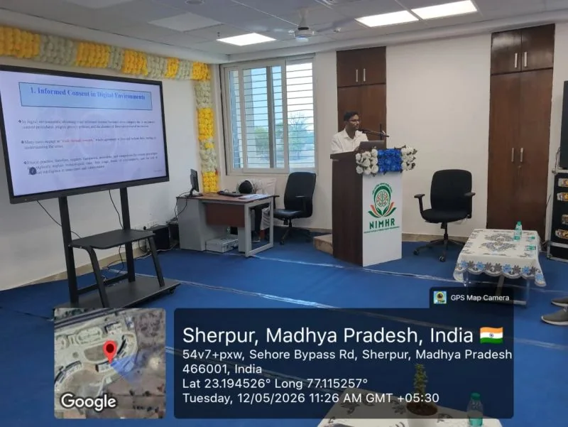 एनआईएमएचआर सभागार में स्क्रीन और पोडियम के पास बोलता वक्ता; तारीख का overlay नीचे